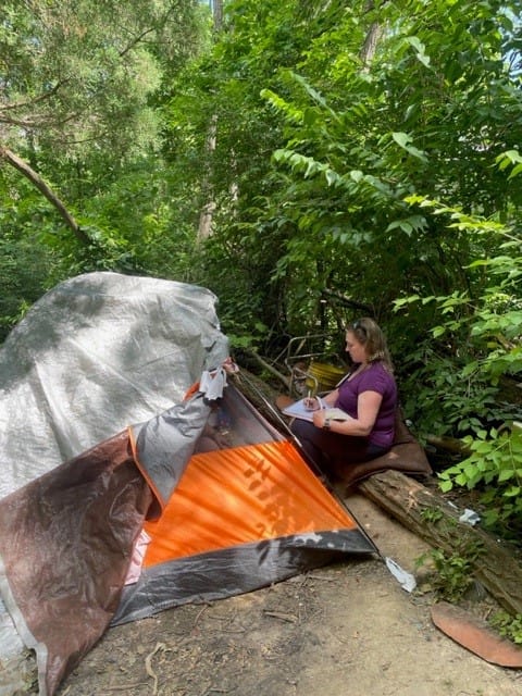 a woman, writing in a notepad, sits on a log outside of a tent in a forested area
