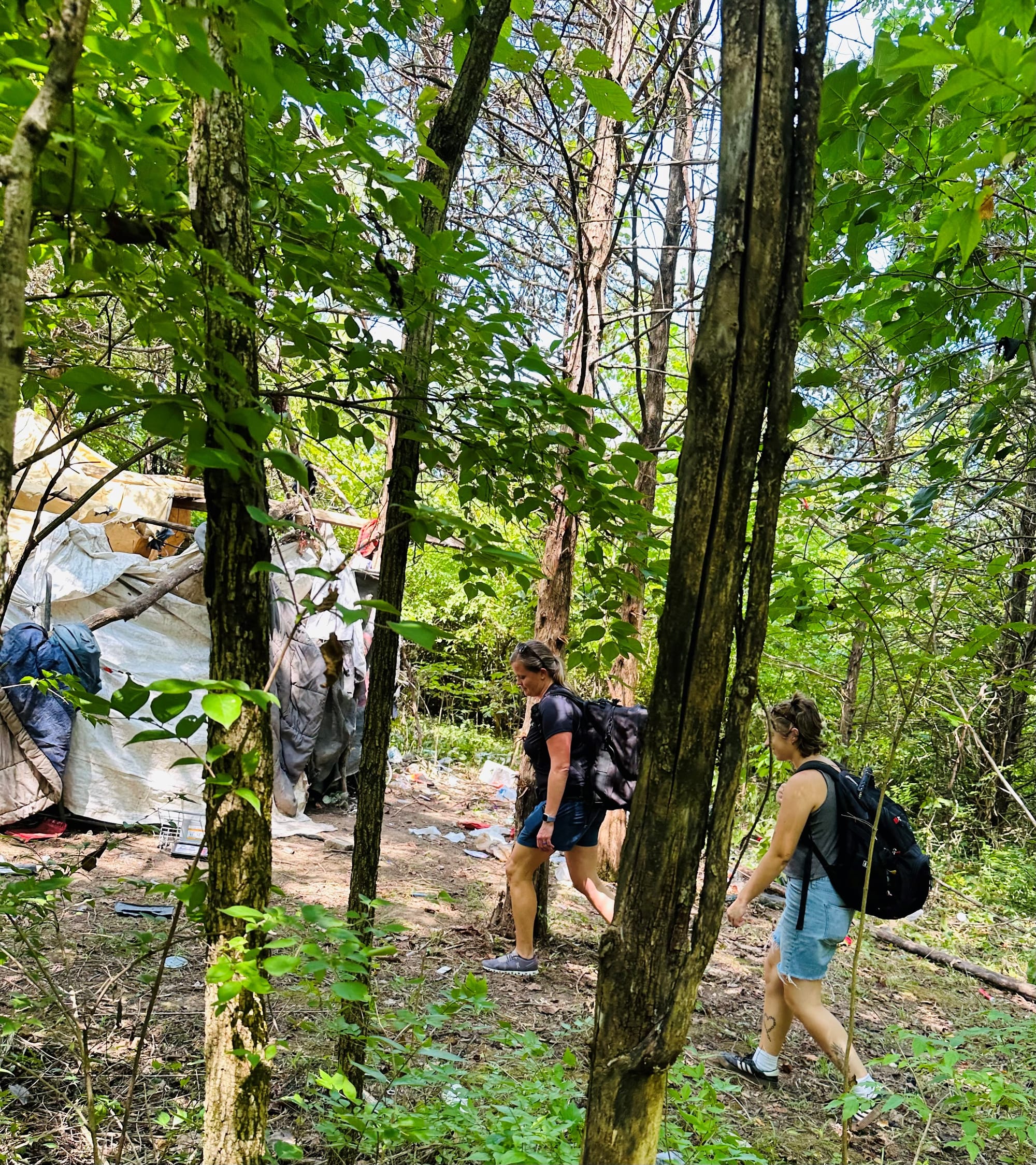 two women with backpacks walk toward a large tarp structure in the woods