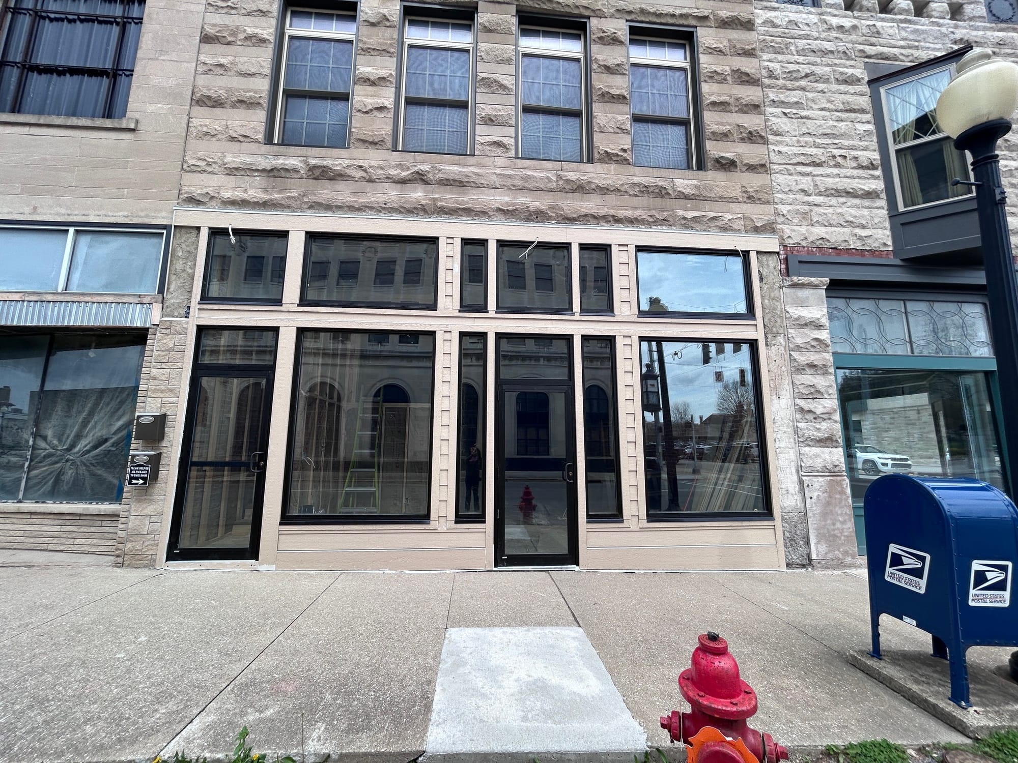 the front of a limestone building with large black-framed glass doors and windows facing the sidewalk. A fire hydrant, blue postal box, and a streetlight appear in the foreground