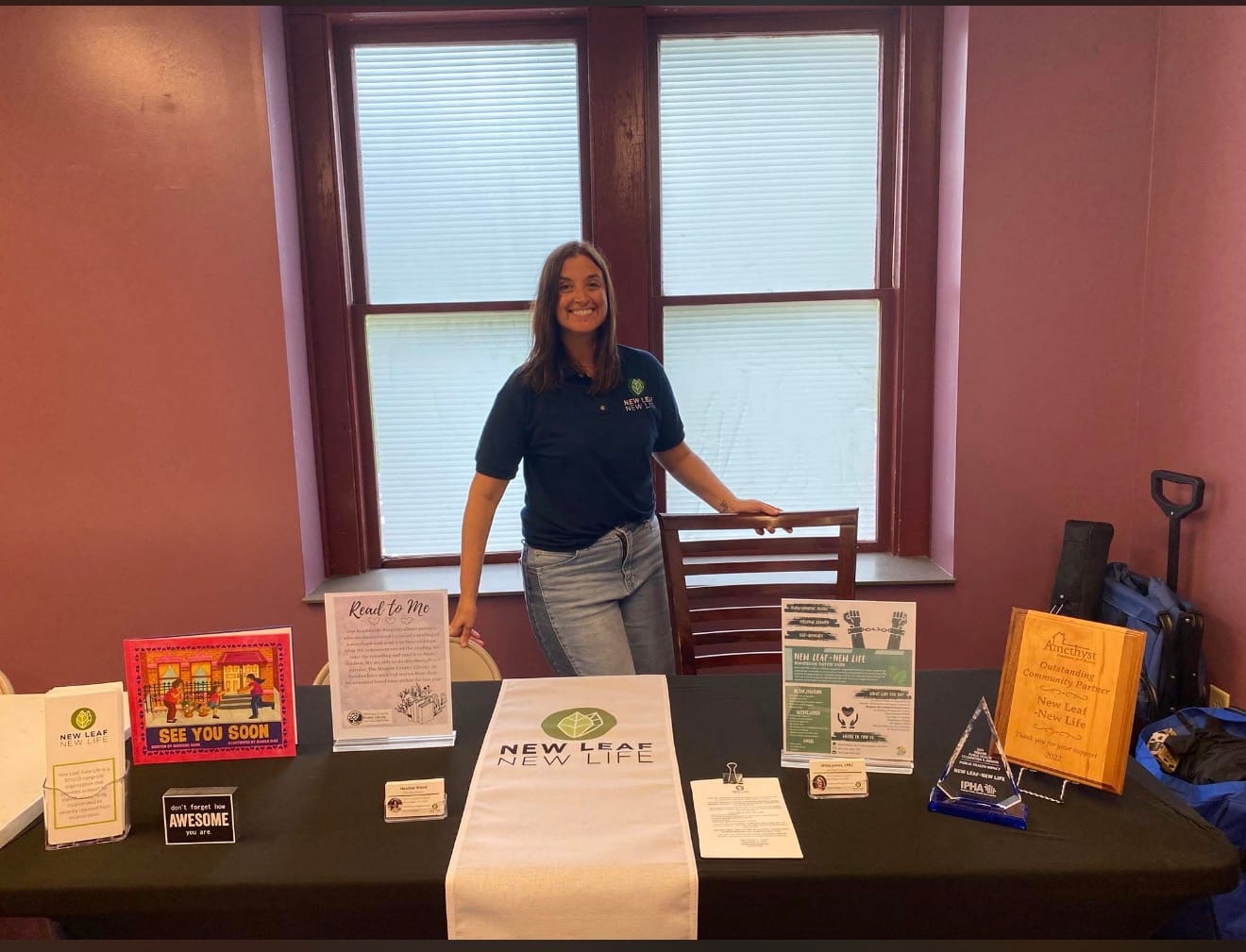 a woman wearing a blue polo and denim jeans, stands behind a table displaying pamphlets and other branded information about New Leaf, New Life