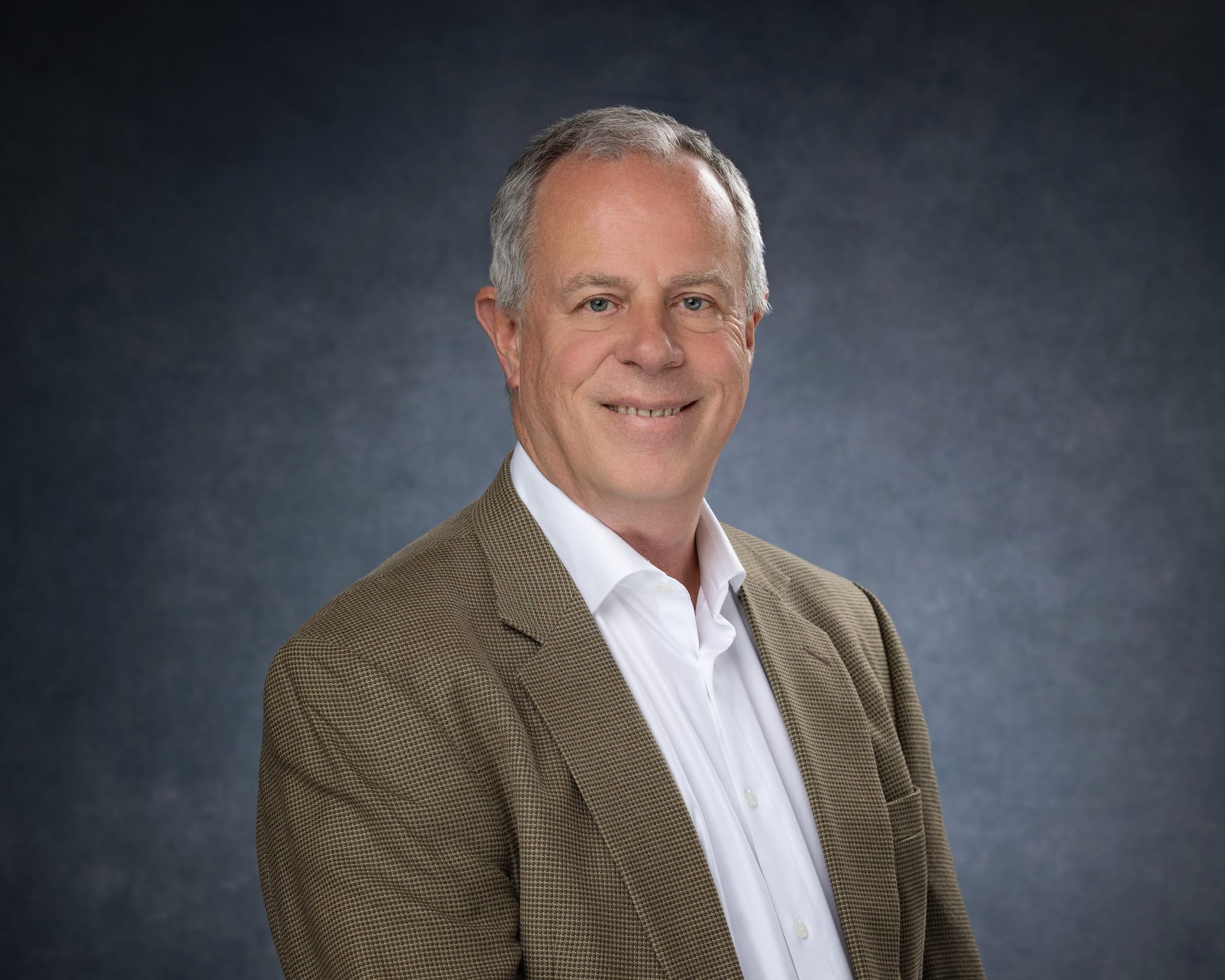 bust portrait of a grey-haired white man wearing a white button-up shirt and brown suit jacket