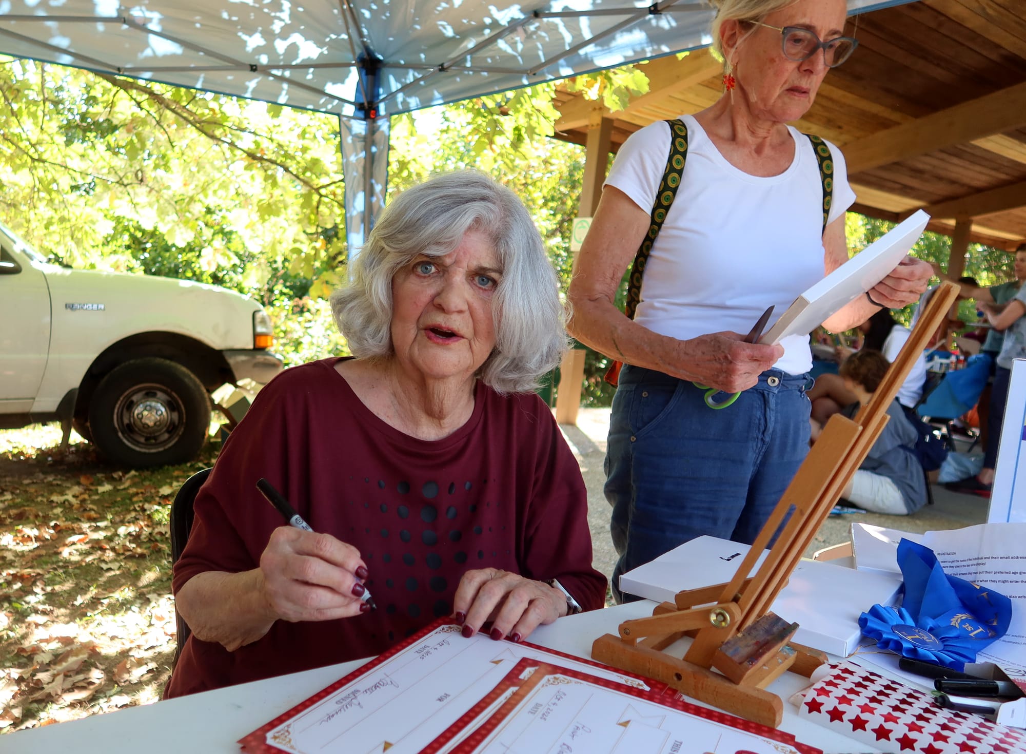 a grey-haired woman with intense blue eyes looks directly into the camera. She's seated at a table under a tent pavilion, signing papers with a Sharpie pen