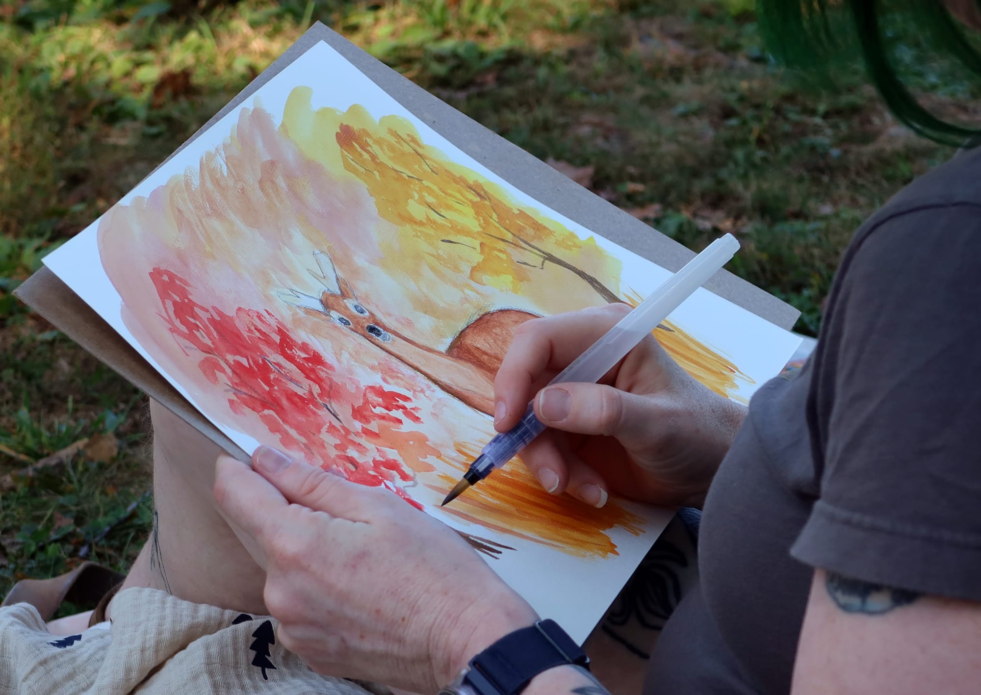 a woman's hands holding a watercolor brush pen and a painting of a deer standing amongst red and yellow trees