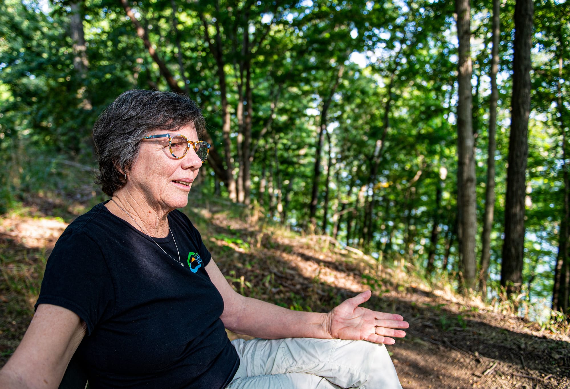 a woman sits on a hillside in the forest, speaking and gesturing