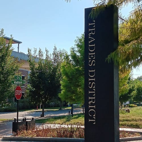 a black column with the words "TRADES DISTRICT" printed sideways along the column's vertical edge. In the background: trees and grass, sidewalks, and a road intersection with a stop sign and road signs for 10th and Madison