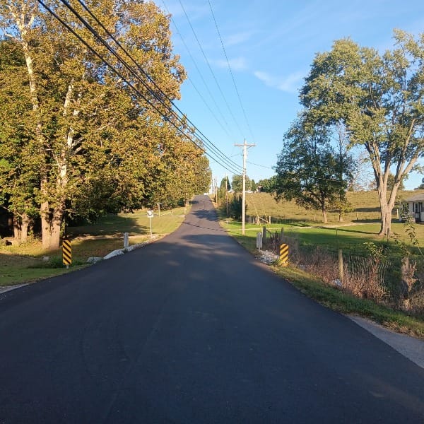 a recently repaved section of rural road, trees, a field of corn, and a house line the road. Powerlines cross overhead. Two black and yellow striped bollards mark a small culvert under the road