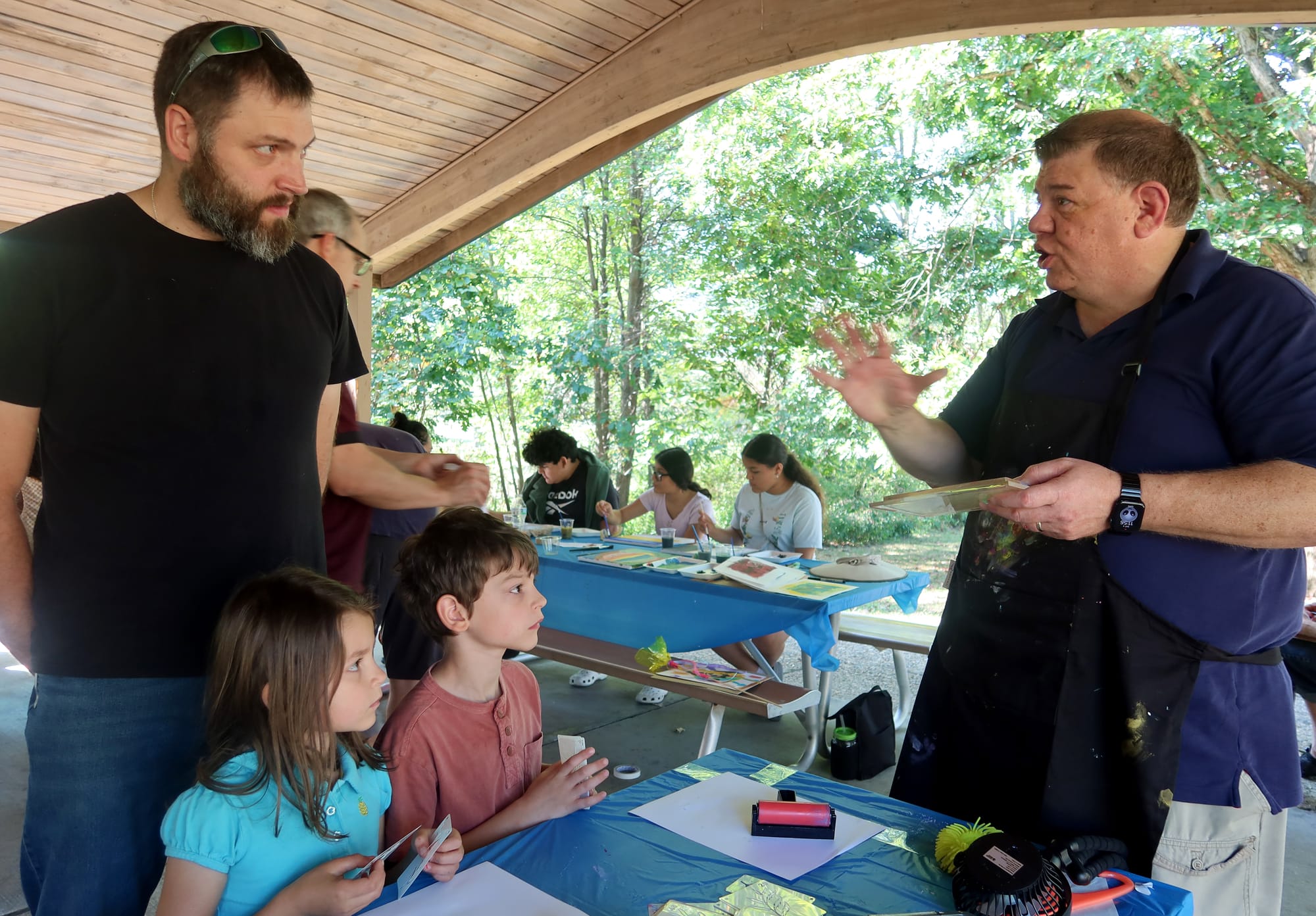 a man in a blue shirt and black apron gestures toward two children and their father as they participate in art activities inside a park shelter house
