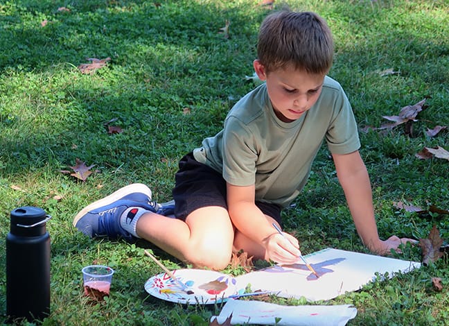a young boy sits in the grass, painting a tree