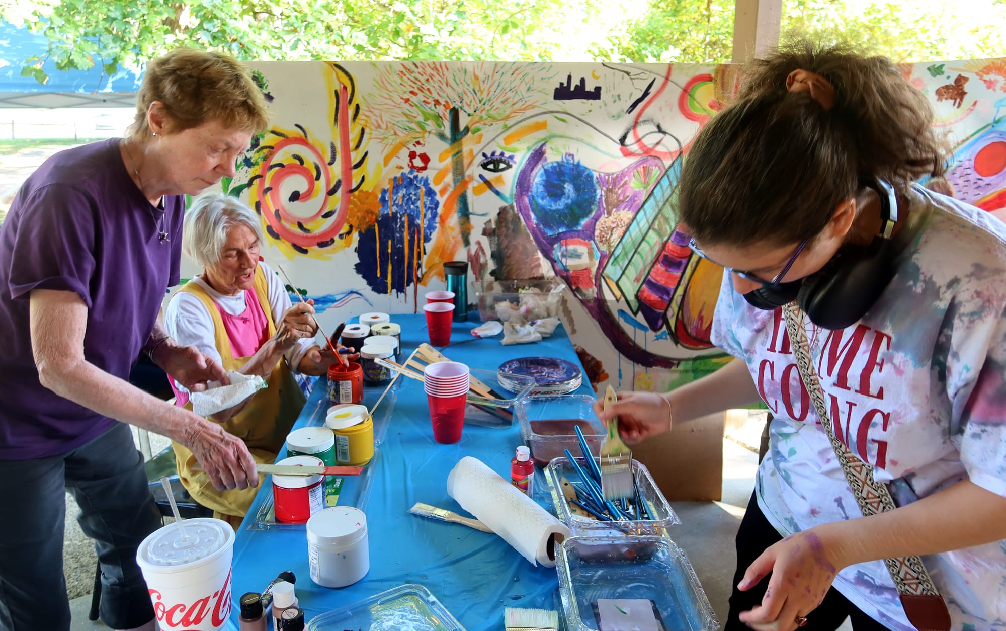 three women at a blue-cloth-covered picnic table share painting supplies as they work on a large mural