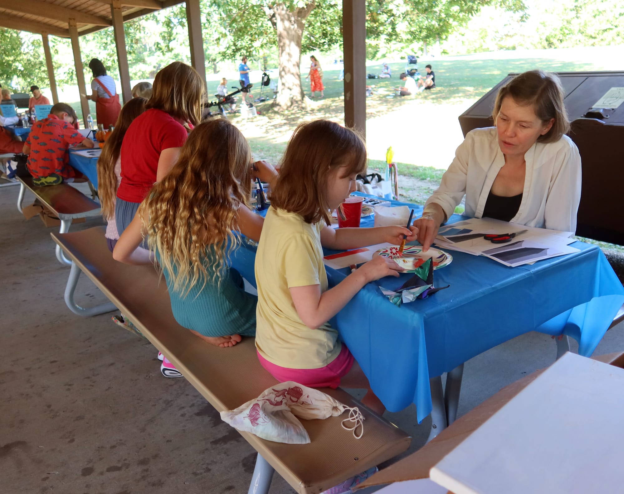 A woman instructs children how to paint. They are seated at a picnic table with a blue plastic tablecloth, covered with art supplies, inside an open-air shelter house at a public park