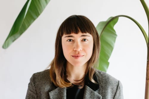 bust portrait of a woman with straight brown hair wearing a grey suit jacket and black shirt. A palm tree contrasts against a white background behind her