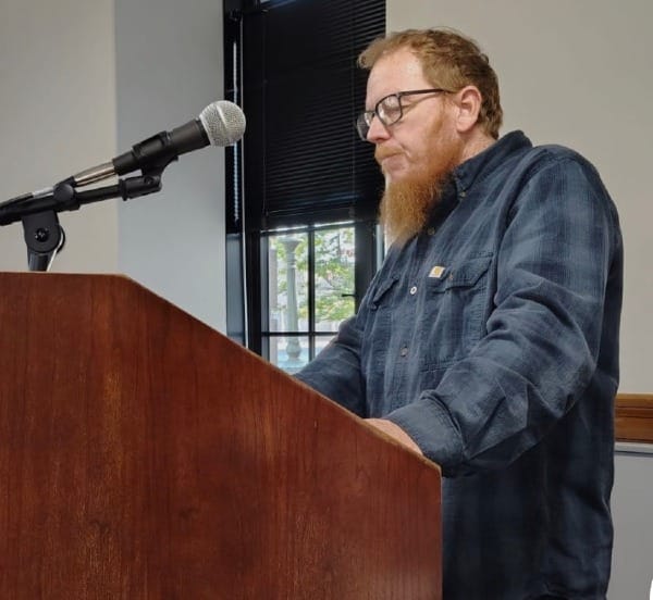 a bearded man wearing black glasses and a blue flannel Carhartt shirt stands at a wooden podium