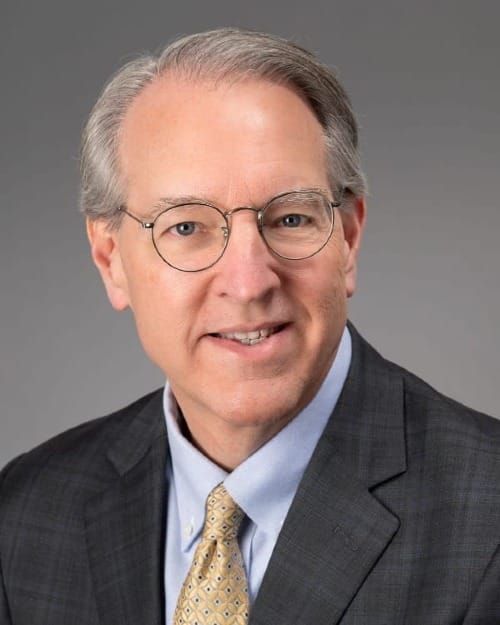 bust portrait of a grey-haired man wearing glasses, a blue button-down shirt, yellow tie, and grey flannel suit jacket