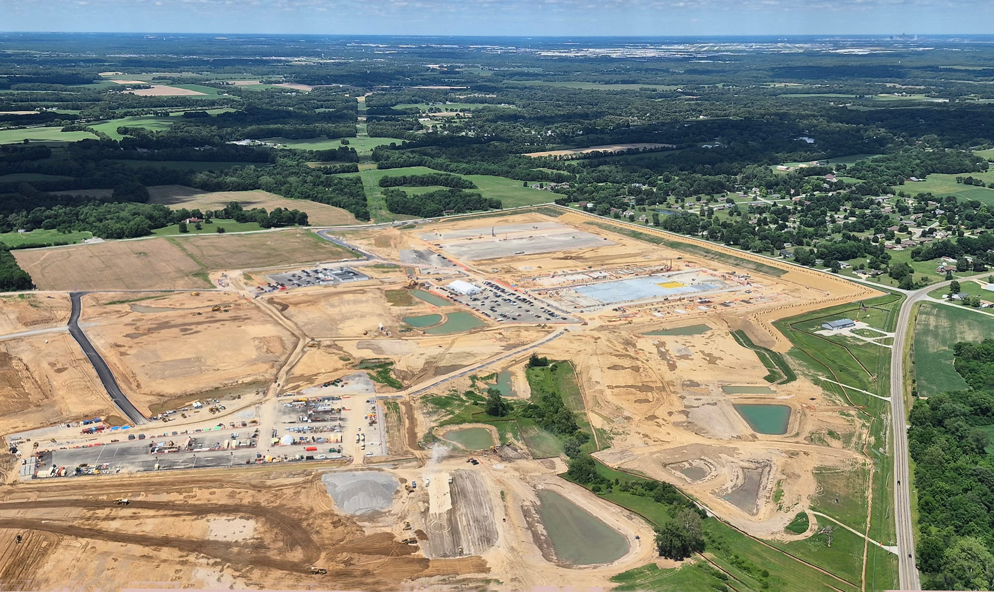 aerial photo of a large construction site in a forested, rural area. Houses and fields are visible under a vast blue sky. Indianapolis skyline on the horizon