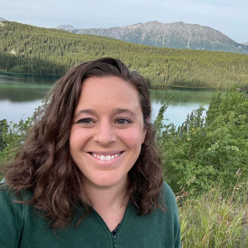 photo of a smiling woman wearing a green sweater. Coniferous forest, a lake, and a mountain in the background