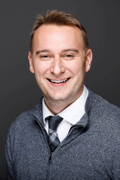 bust portrait of a blond man wearing a white button-up, grey tie, and grey zippered sweater