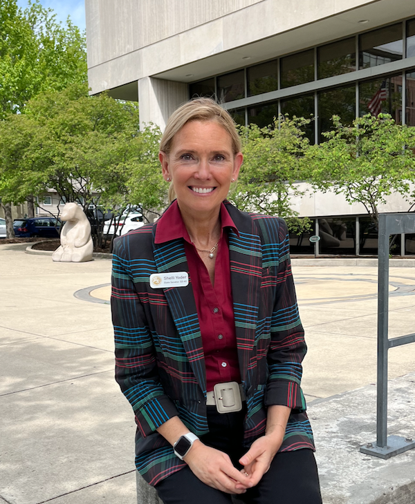 photo of a blonde woman sitting in front of the Monroe County Public Library. She's wearing a red button-up shirt and multi-colored blazer