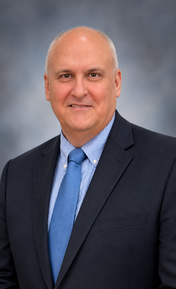 bust portrait of a man, mostly bald, with short grey hair, dark brown eyes, and a blue and white striped button-down shirt and blue tie under a navy blazer