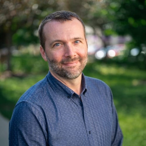 bust portrait of a man in a blue button-up shirt. Blurry bushes and trees make up the background