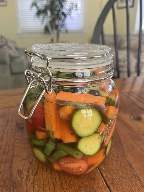 a glass jar with sealable lid, filled with cut carrots, cucumber, tomatoes, and green beans