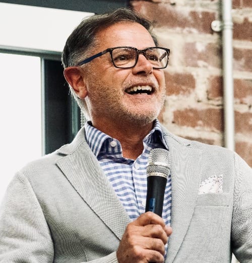 A man wearing dark glasses, a light grey suit jacket, and white and blue button-up, holding a microphone, standing in front of a brick wall