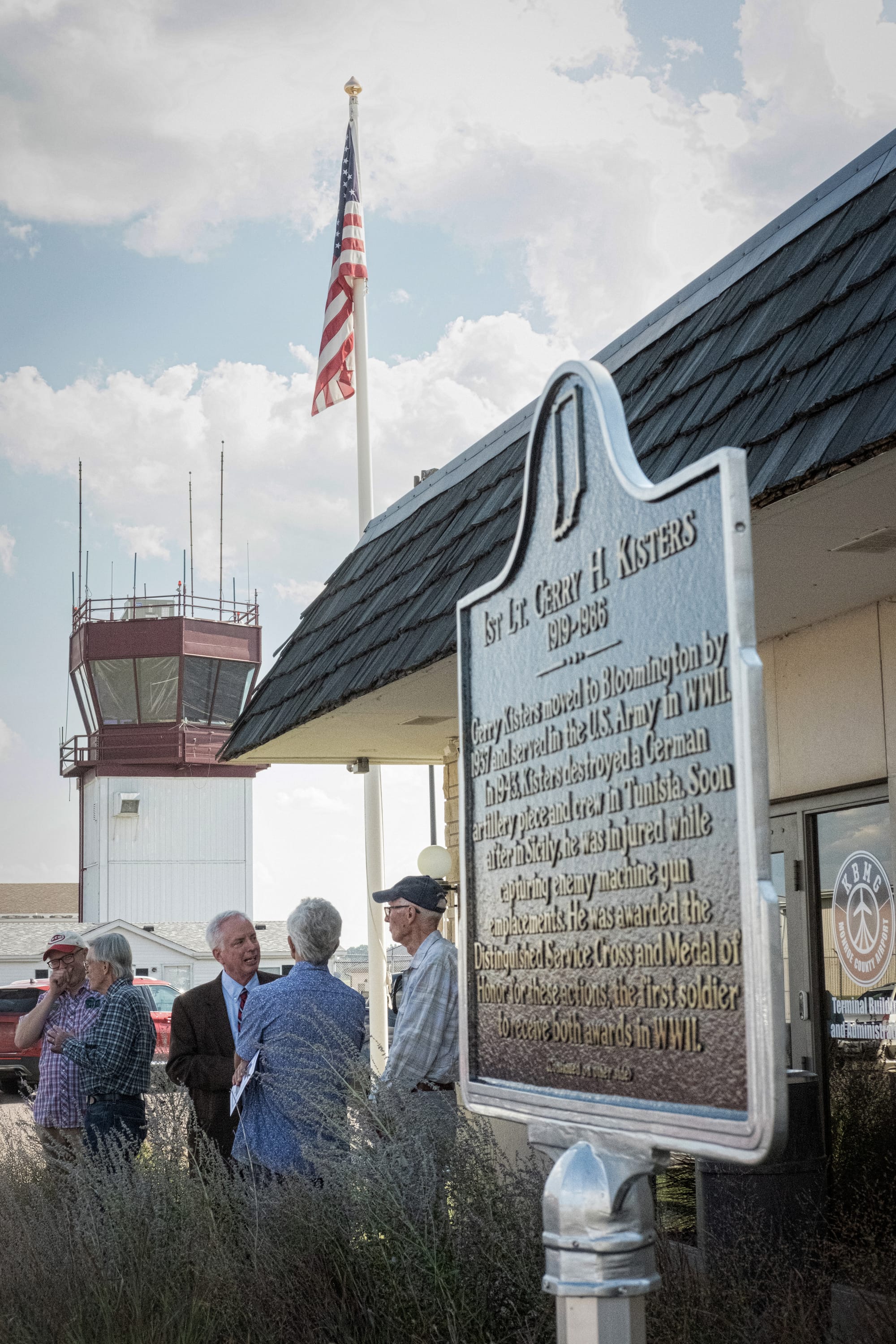 five men stand around outside the airport buildings near the Kisters historical marker