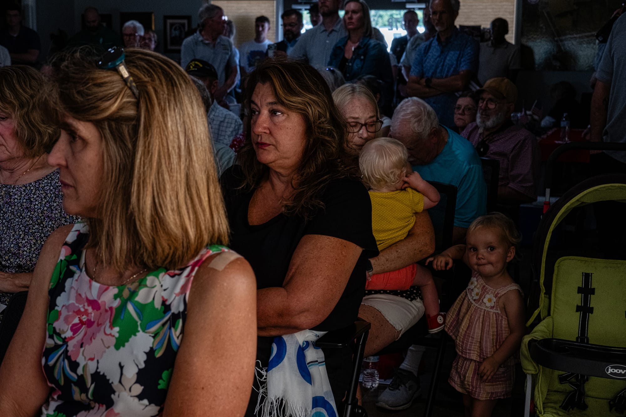 a room full of people, some sitting, some standing. A cute baby girl looks directly at the camera