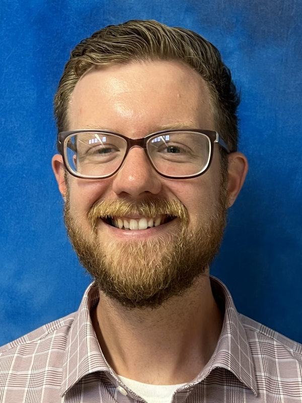 portrait of a smiling, bearded man against a blue background. He wears dark-rimmed glasses and a white and red checkered shirt