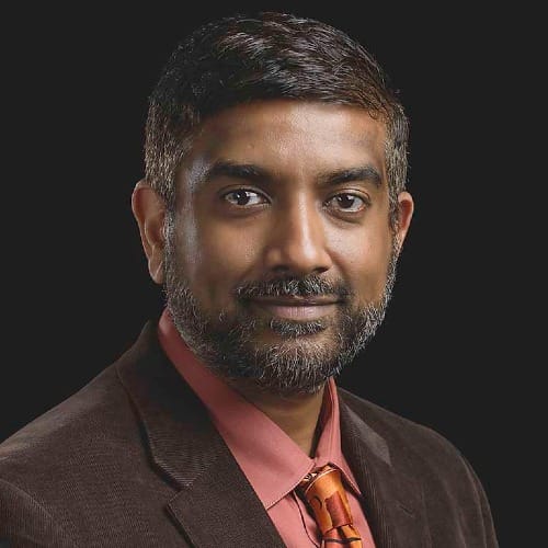 headshot of a man with a short beard. He wears a salmon-colored button up shirt, orange tie, and brown suit jacket.