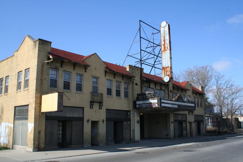 the dilapidated exterior of a Spanish Mission-style theater building, with yellow brick walls and a red roof. The marquee is blank except for a website: rivolitheatre.org. The large, blue, vertical sign depicting the theatre's name is rusted