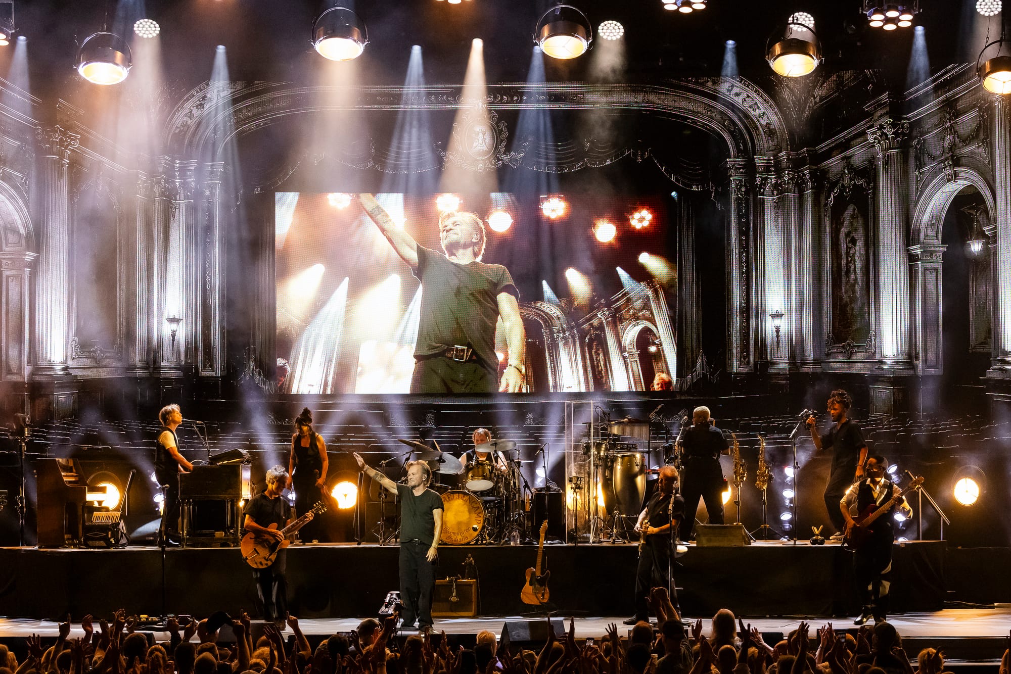 John Mellencamp, wearing a black t-shirt and black pants, waves to the crowd from onstage, surrounded by yellow stagelights and an eight-piece band, in a large, ornate, indoor amphitheater
