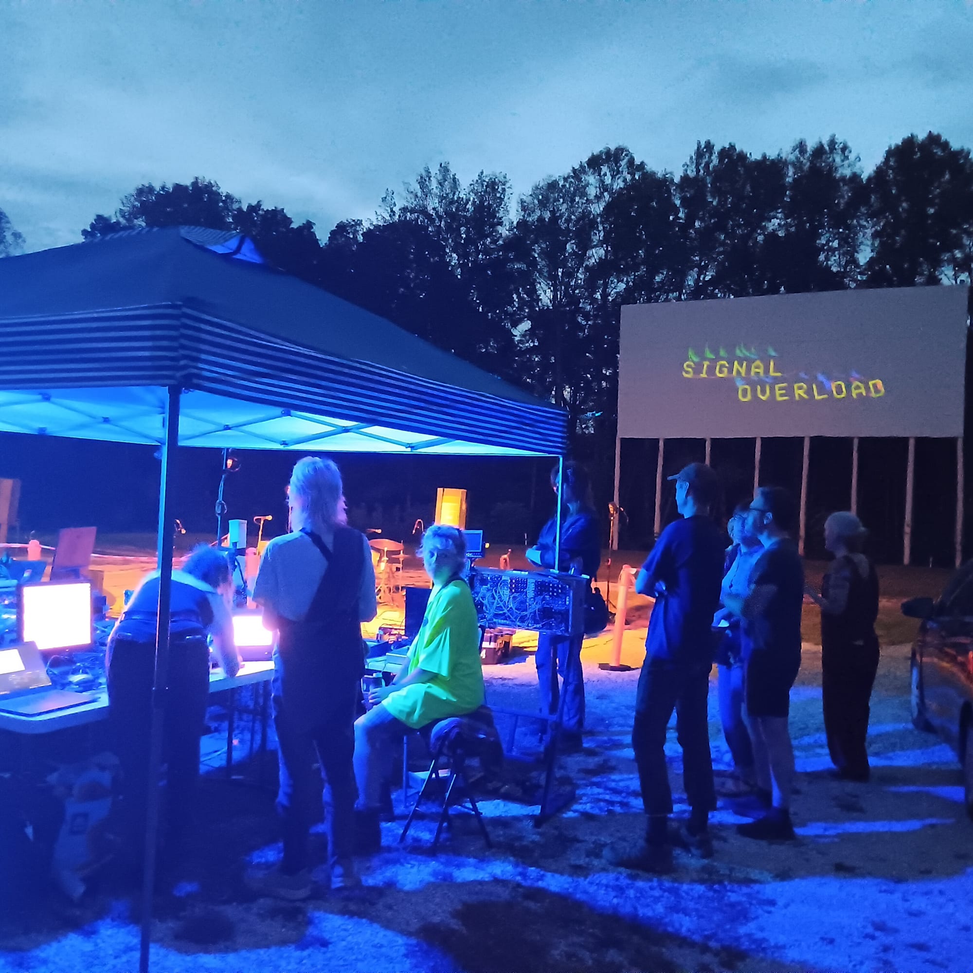 a crowd of people gathers near a tent filled with screens and audio equipment. In the background, a drive-in theater screen with the words "SIGNAL OVERLOAD" in yellow