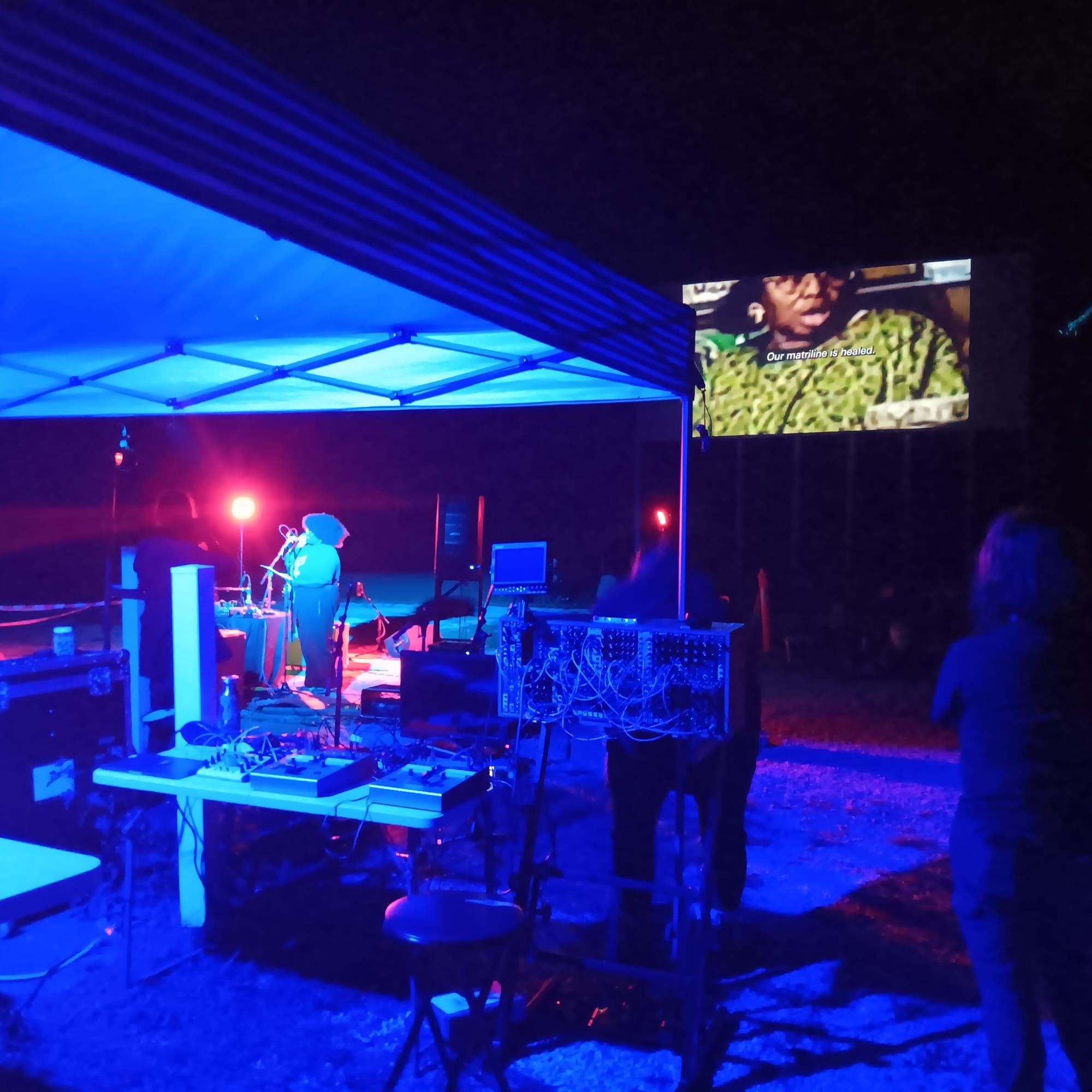a Black woman, lit by blue light, stands at a microphone, surrounded by sound equipment. A drive-in theater screen behind her depicts an older Black woman and the words "Our matriline is healed."