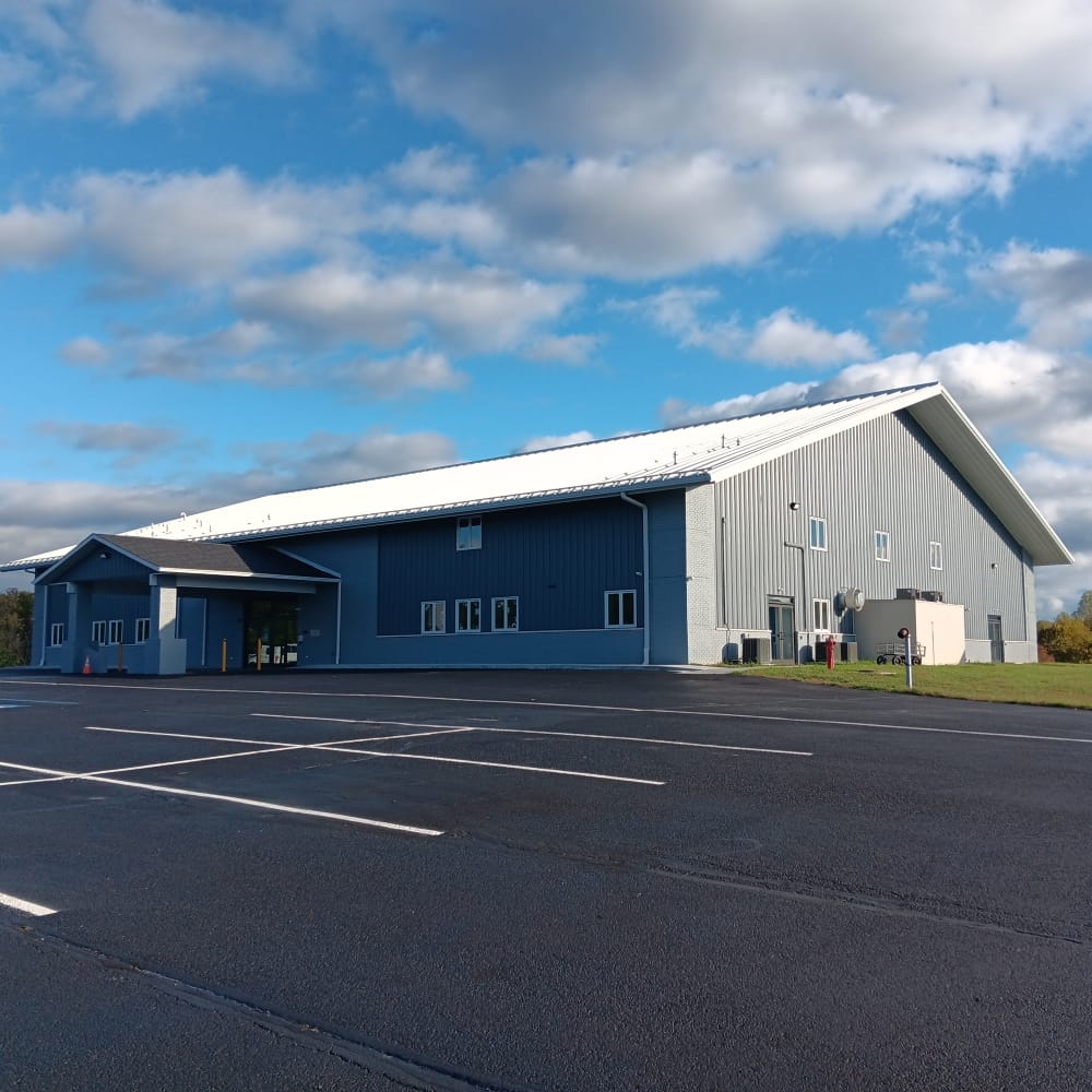 a large grey and white metal building contrasting against a black asphalt parking lot and a blue sky full of billowing white clouds