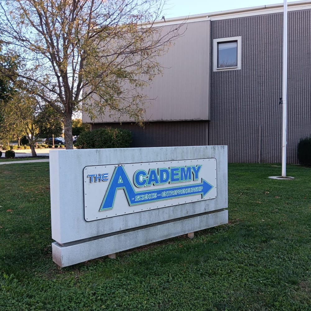 a stone sign with blue and green lettering for "The Academy of Science and Entrepreneurship." The school building, made of corrugated cement and brown metal siding, and one square window, are visible in the background, as well as a flagpole and some trees 