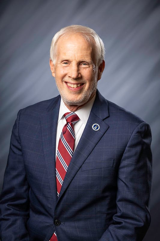 a white-haired man wearing a white button-down shirt, red, white, and navy striped tie, and navy flannel suit jacket