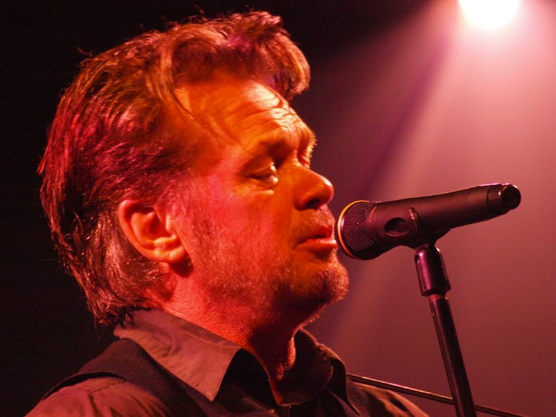 A close up of John Mellencamp, lit by red theater lights, singing into a microphone
