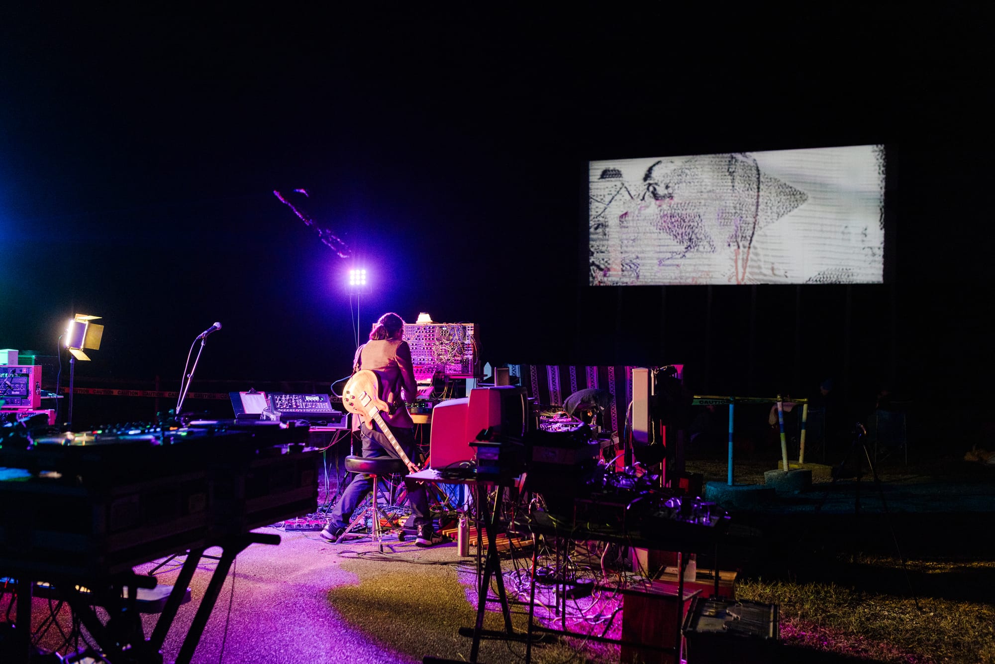 a man in a brown suit jacket has a guitar strapped to his back as he controls a synthesizer board. The drive-in screen displays a white screen with black markings that somewhat resembles scribbles on a notebook page