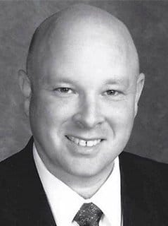 black and white photo of a bald man in a black suit jacket, white shirt, and tie