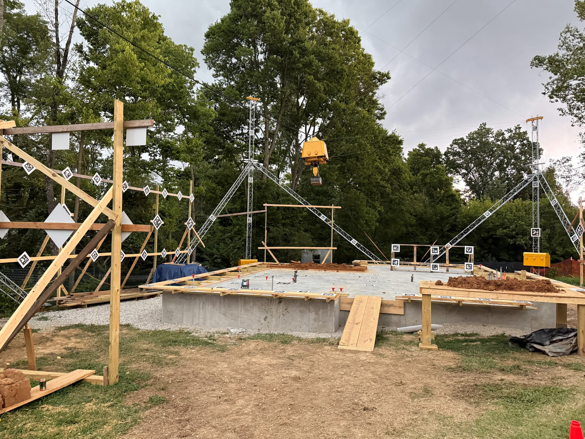 wide angle shot of a home's concrete slab foundation; wooden beams and metal scaffolding surround the base, providing a structured framework for the yellow robot suspended from cables above