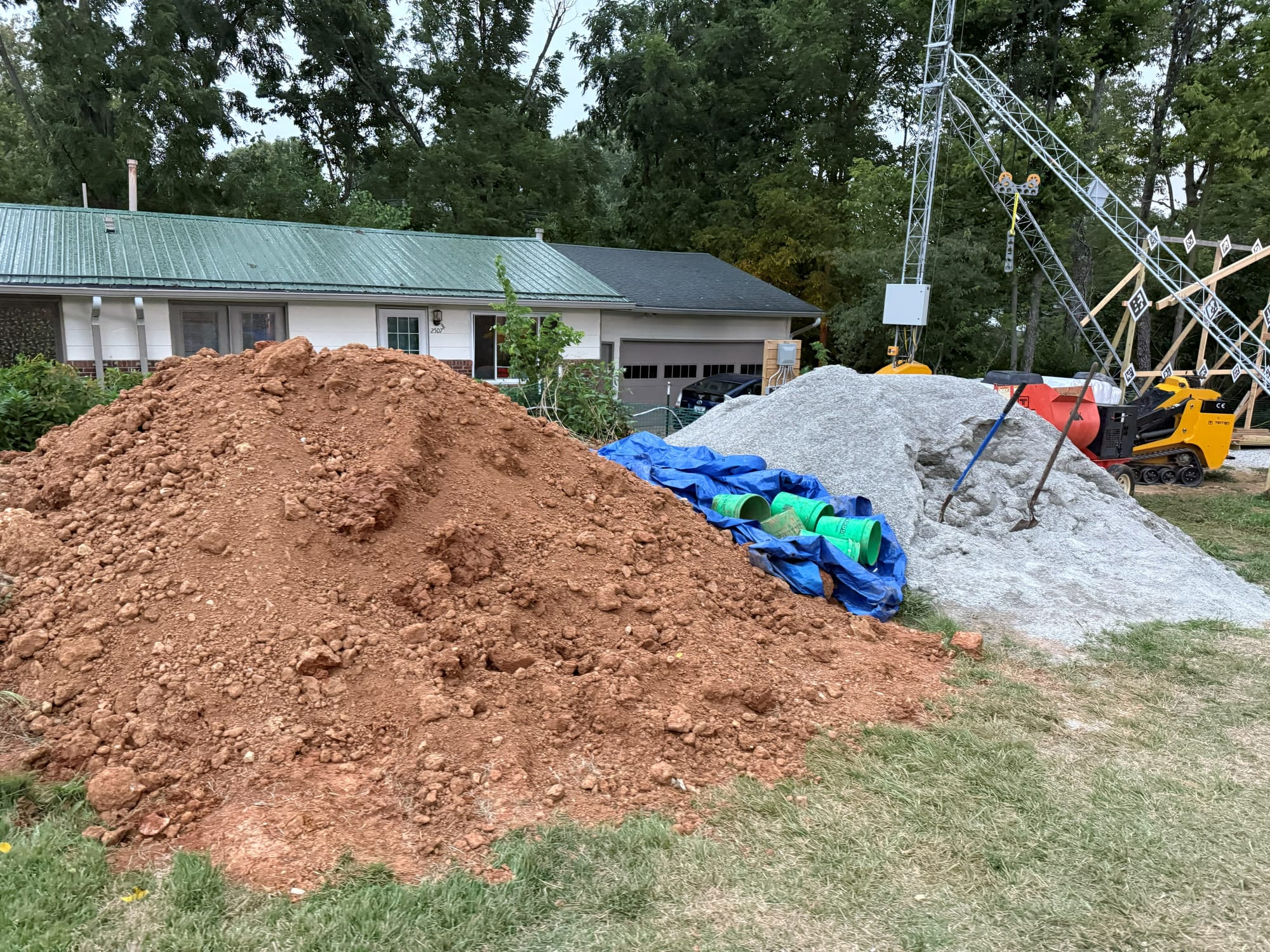 green 5-gallon buckets are piled on top of a blue tarp which is roughly folded between a large pile of clay and a large pile of sand