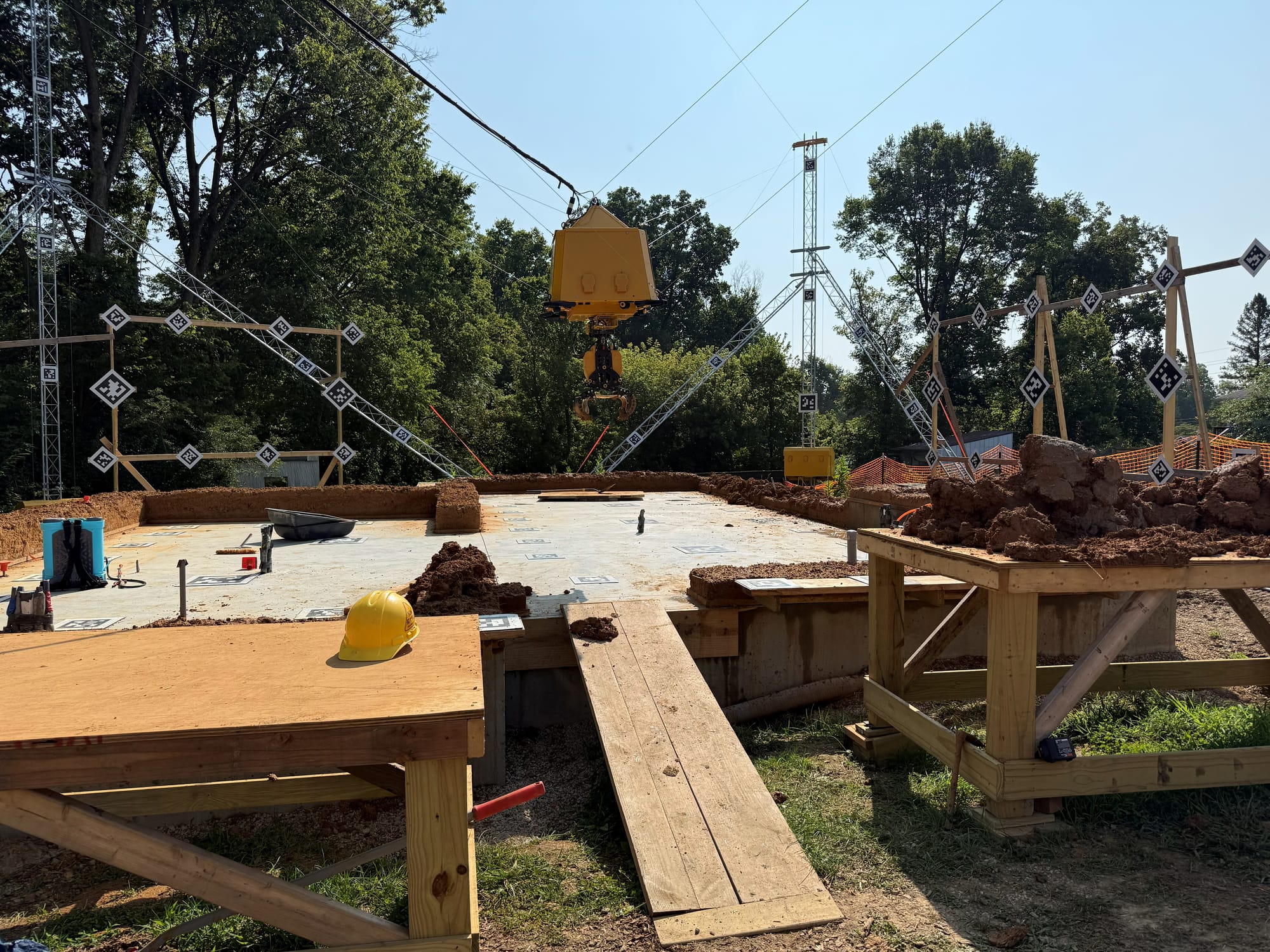 a construction site with wooden tables, a wooden ramp up to a concrete slab, and piles of clay. A yellow robot claw suspended from cables between metal scaffolding hangs above the site
