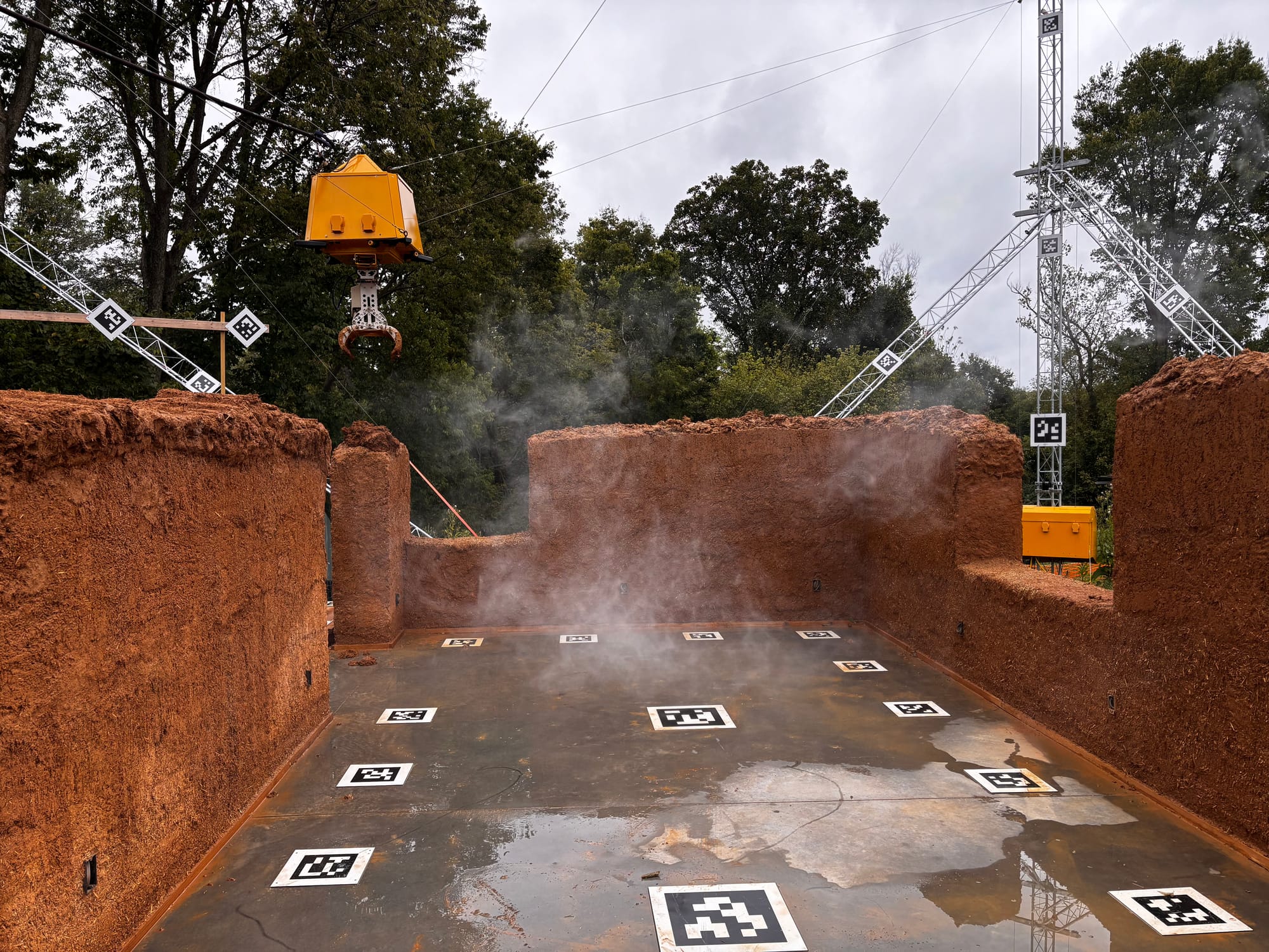 steam rises from a rain-soaked cement floor surrounded by cob walls. A yellow robotic claw hangs suspended from cables above