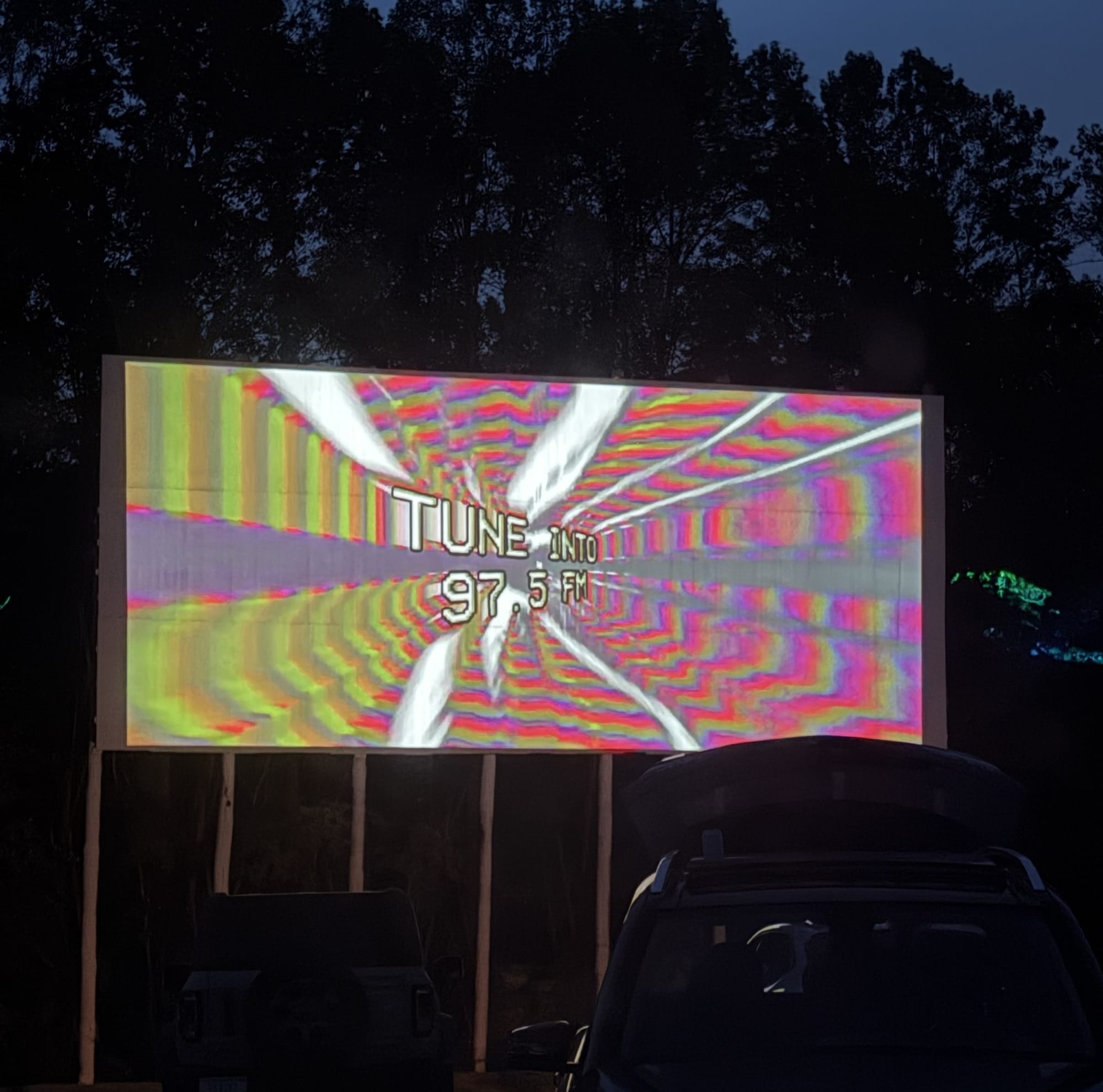 a drive-in theater screen showing a psychedelic burst of red, yellow, white, and grey with the words "TUNE INTO 97.5 FM"
