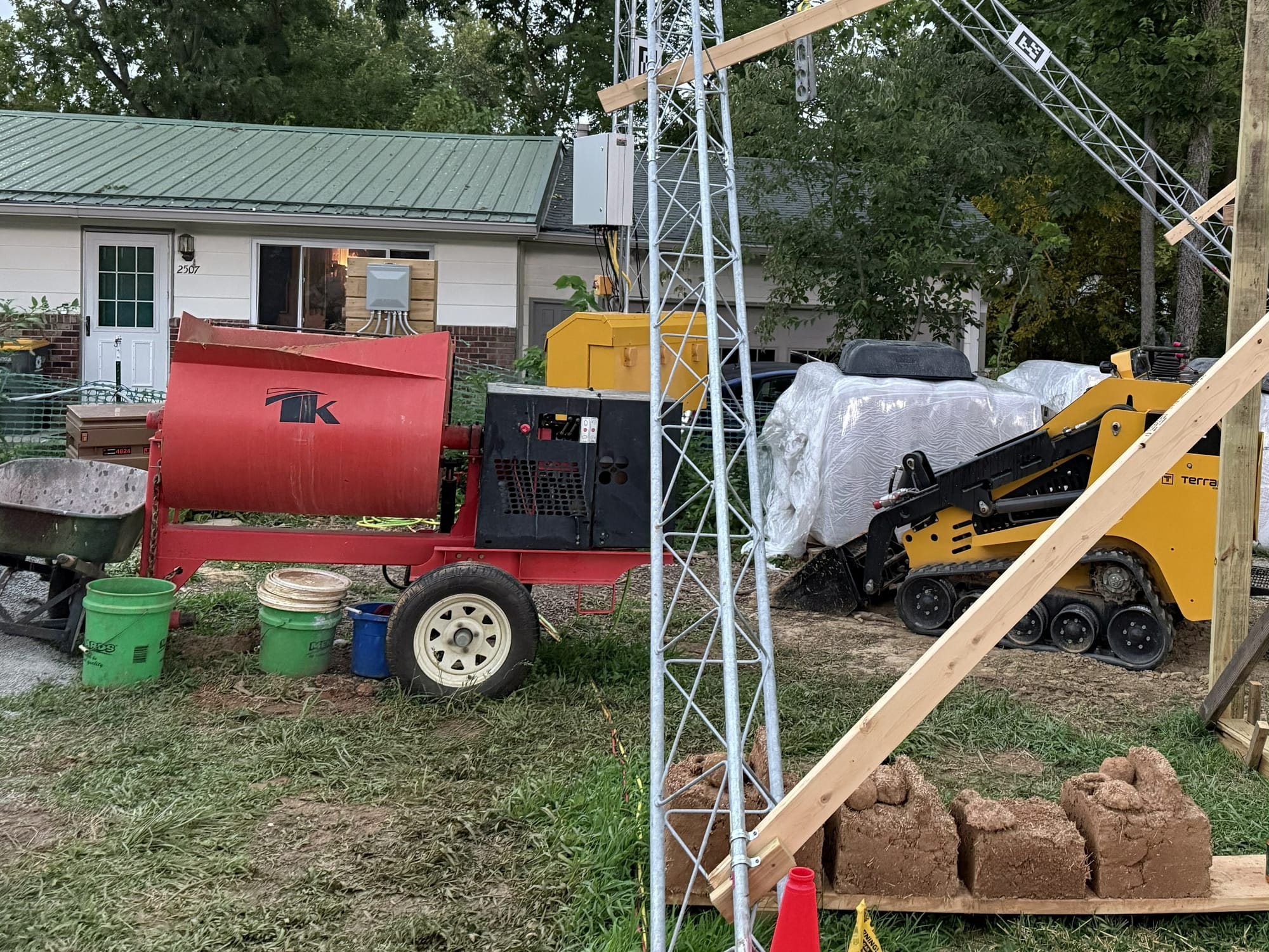 a wheelbarrow sits next to a red machine mixer; a small yellow front-loader is parked in the dirt nearby. Four cob bricks sit on a plank of wood in the foreground.