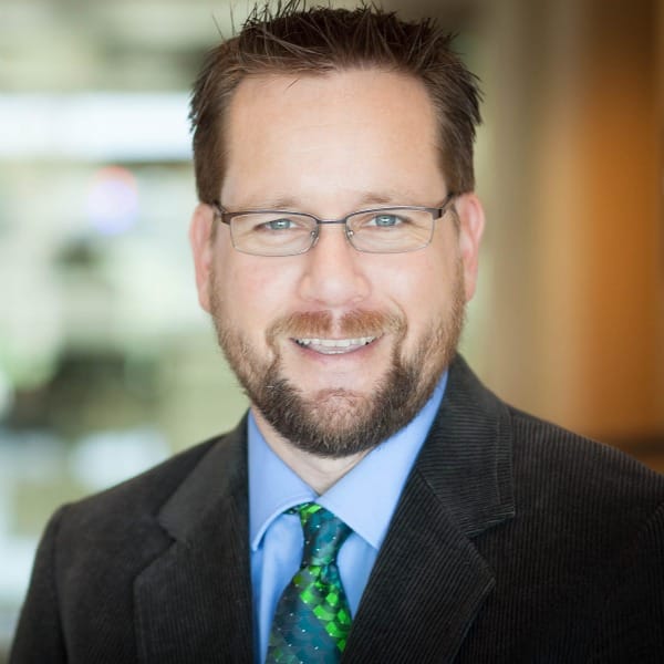a brown-haired man with narrow glasses and a beard. He wears a blue button-up shirt, blue and green tie, and brown corduroy jacket