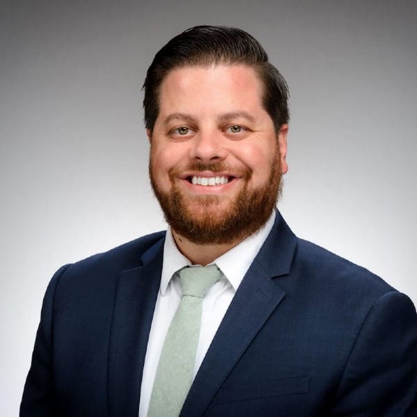 a brown-haired bearded man wearing a white button-up shirt, grey tie, and navy suit jacket