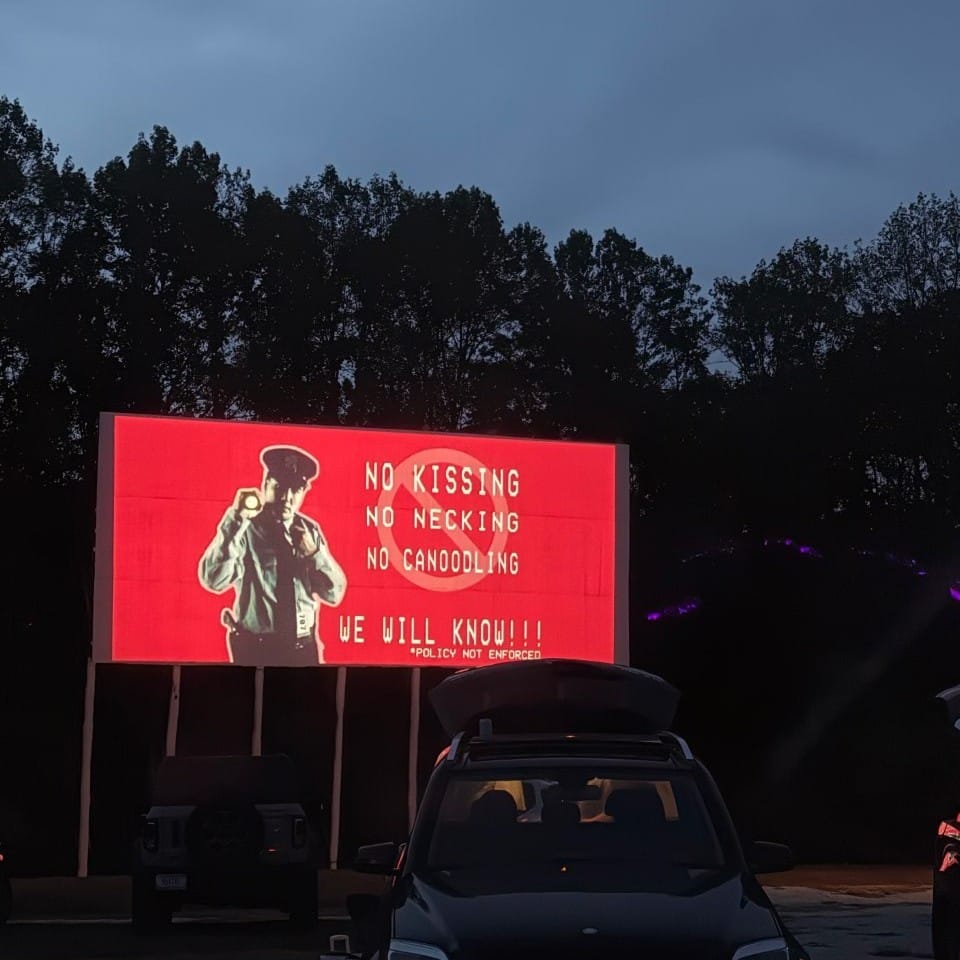 a drive-in theater screen depicting a police officer against a bright red background with the words “No Kissing, No Necking, No Canoodling. We Will Know (policy not enforced).”