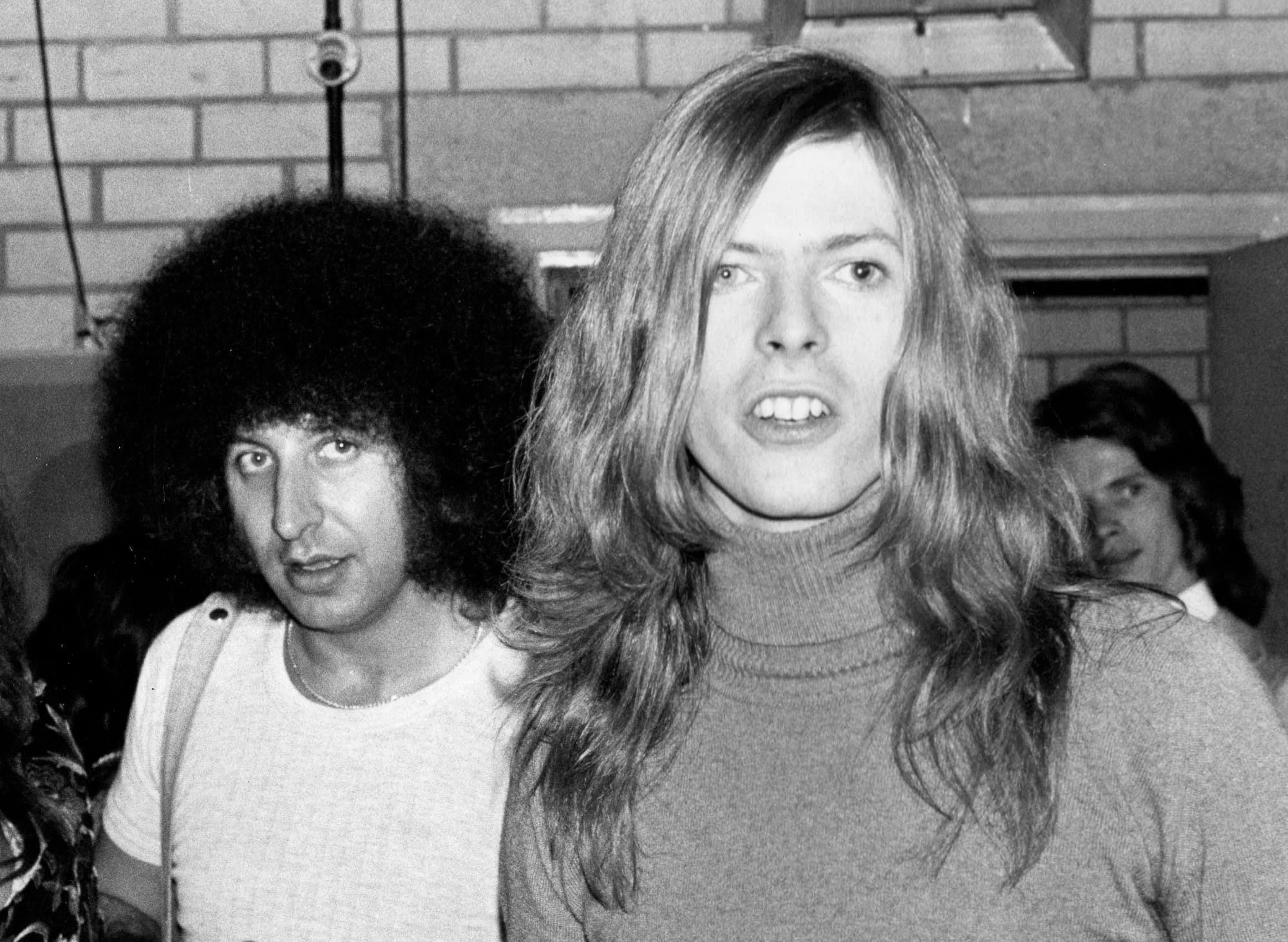 Tony DeFries, a man with a large afro and wearing a white shirt, beside David Bowie, wearing a grey turtleneck sweater, his face framed by long blond hair