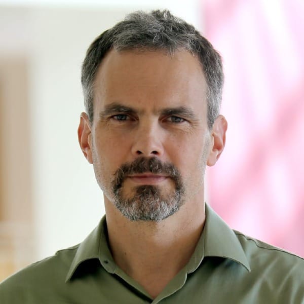 a salt-and-pepper haired man with dark eyes and a goatee wearing a green button-up shirt